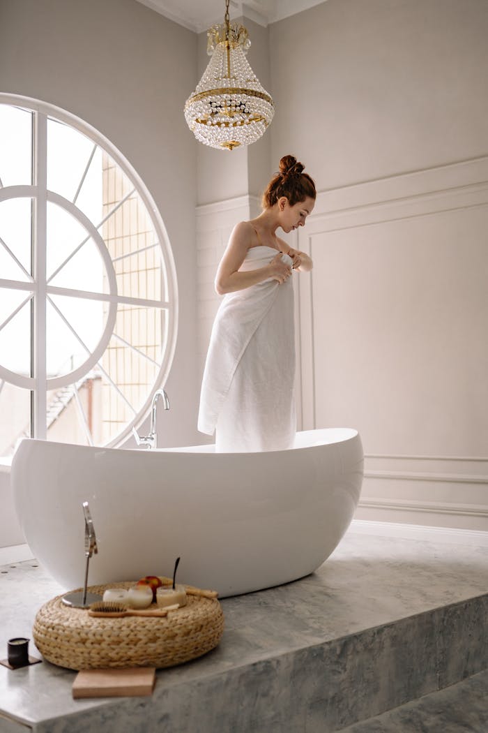 A woman stands in a luxury bathtub, wrapped in a towel, in a bright bathroom.