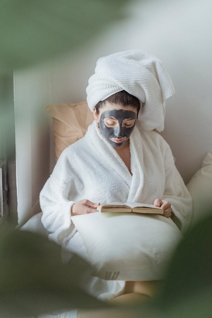 Relaxing self-care moment with a woman in a robe, clay mask, and book, offering tranquility and pampering.